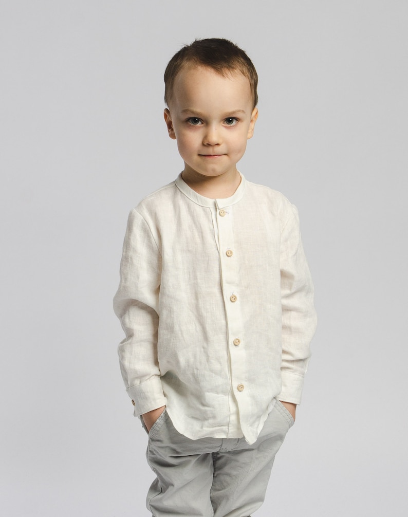 May include: A young boy wearing a white linen shirt with a mandarin collar and grey pants. He is standing with his hands in his pockets and looking directly at the camera.