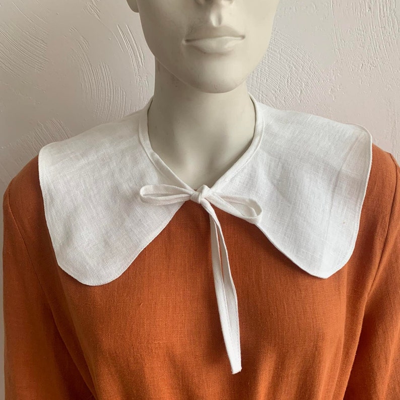 May include: A white linen collar with a bow tie, worn over a rust-coloured dress.