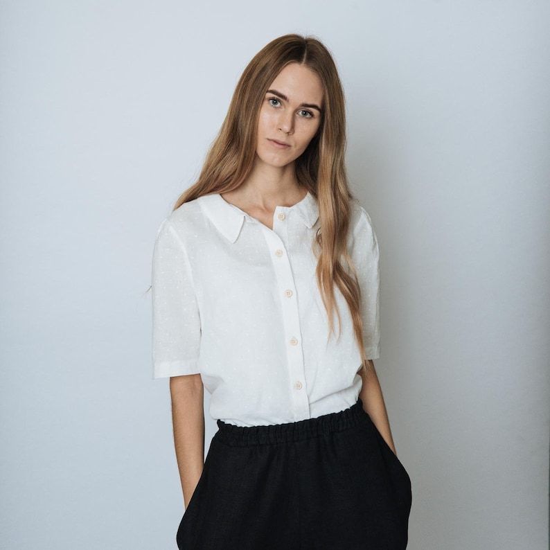 May include: A woman wearing a white collared shirt with a button-up front and black trousers.