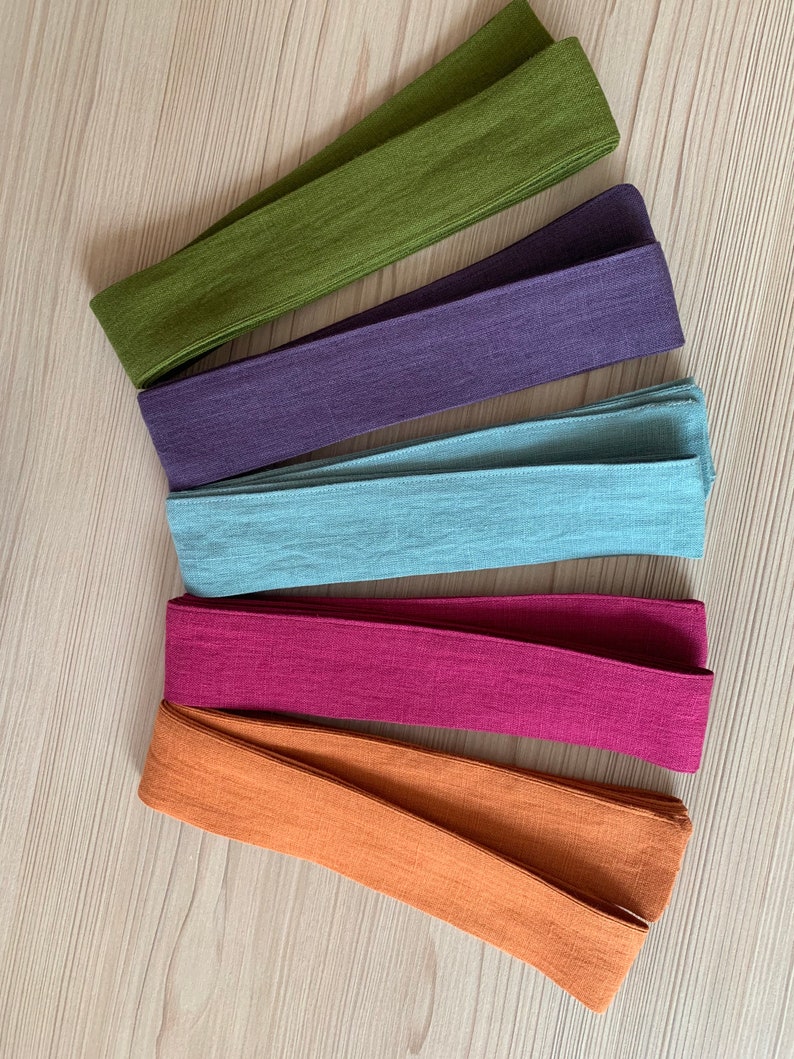 May include: Five fabric ribbon strips in different colors: olive green, purple, light blue, burgundy, and orange. The strips are arranged in a stack, with the olive green strip on top and the orange strip on the bottom.