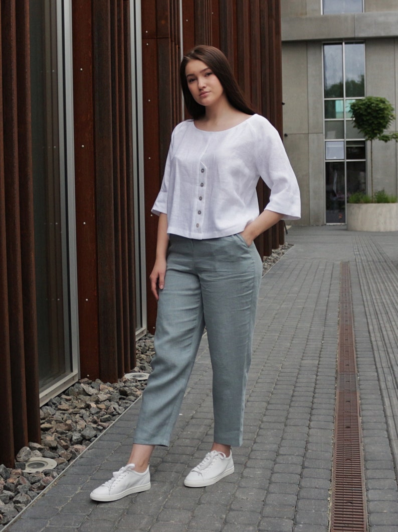 May include: A woman wearing a white linen top with a button-up front and a pair of light blue linen pants. She is wearing white sneakers and has her hand in her pocket.