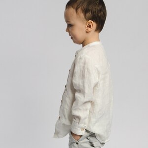 May include: A young boy wearing a white linen shirt and gray pants stands facing away from the camera. He is looking down and has his hand in his pocket.