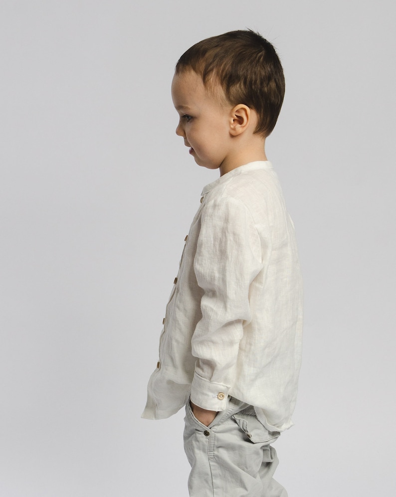 May include: A young boy wearing a white linen shirt and gray pants stands facing away from the camera. He is looking down and has his hand in his pocket.