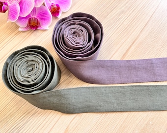 Könnte beinhalten: Zwei Stoffrollen in Grau- und Lilatönen, wobei sich von jeder Rolle ein Stoffstück erstreckt. Im Hintergrund sind rosa Orchideen zu sehen. Der Stoff scheint Leinen oder ein ähnliches Material zu sein.