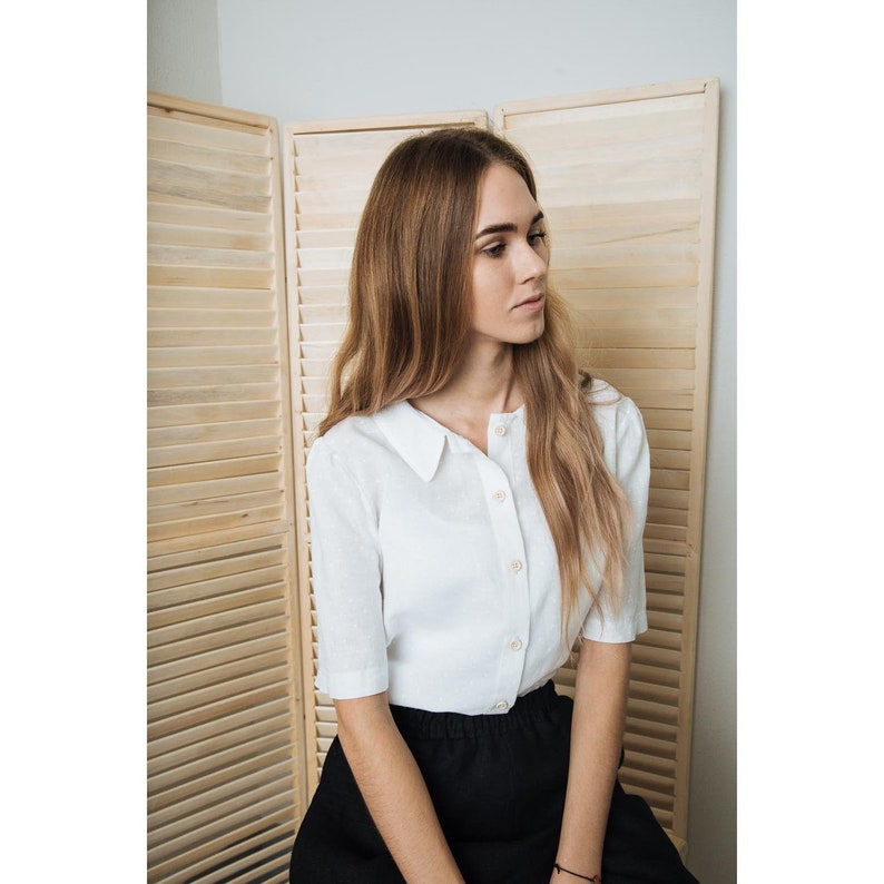 May include: A woman wearing a white linen shirt with a collared neckline and a black skirt. The shirt has a button-up front and short sleeves. The woman is sitting in front of a wooden slat wall.