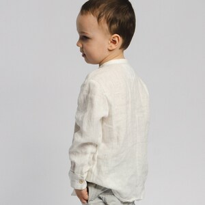 May include: A young boy wearing a white linen shirt and gray pants stands facing away from the camera. He is looking over his shoulder.