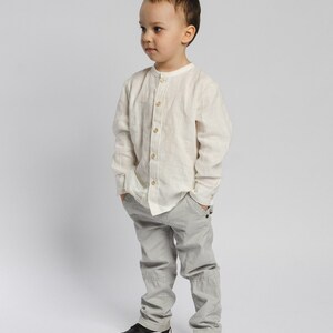 May include: A young boy wearing a white collared shirt and gray pants. He is standing with his hands in his pockets and looking to the side.