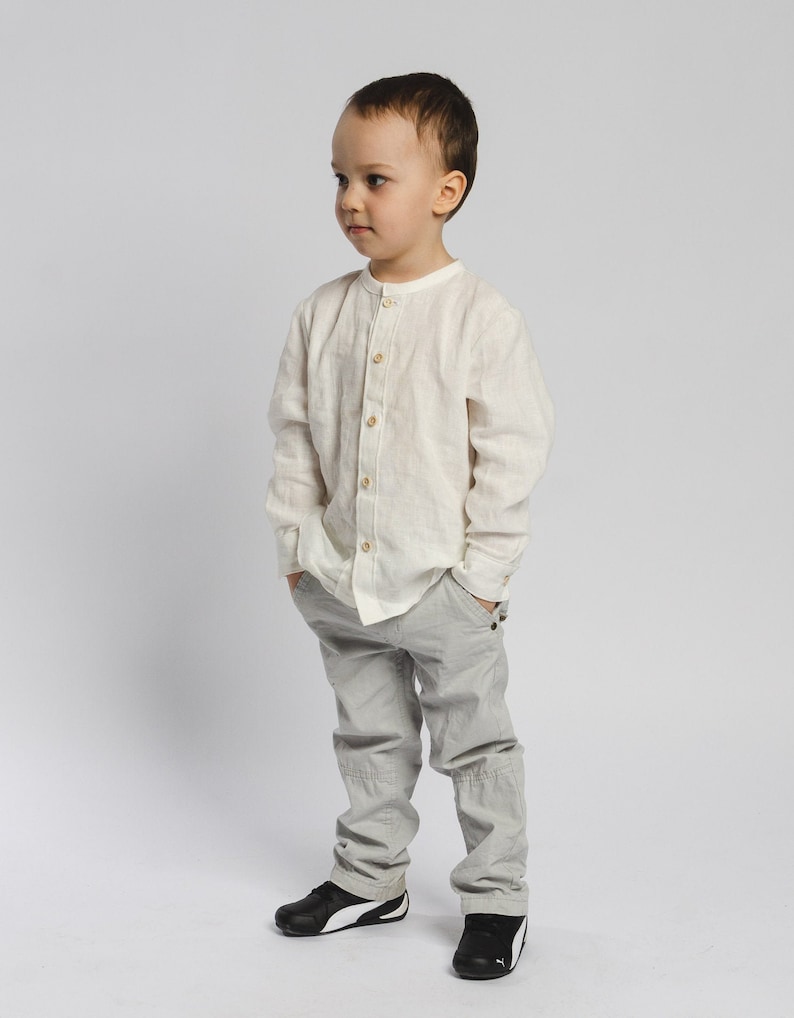 May include: A young boy wearing a white collared shirt and gray pants. He is standing with his hands in his pockets and looking to the side.