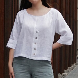 May include: A white linen top with a button-up front and three-quarter sleeves. The top has a relaxed fit and a slightly cropped hem.