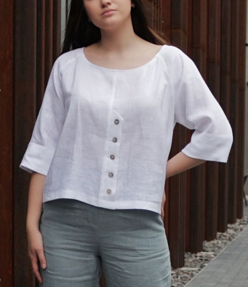 May include: A white linen top with a button-up front and three-quarter sleeves. The top has a relaxed fit and a slightly cropped hem.