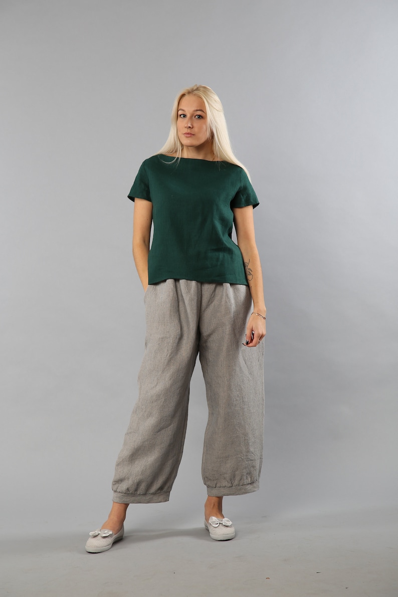 May include: A woman is wearing a dark green linen top with short sleeves and gray linen pants. She is standing on a gray background.