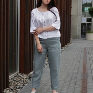May include: A woman wearing a white linen top with a button closure and light blue linen pants. She is standing in front of a brick wall and is wearing white sneakers.