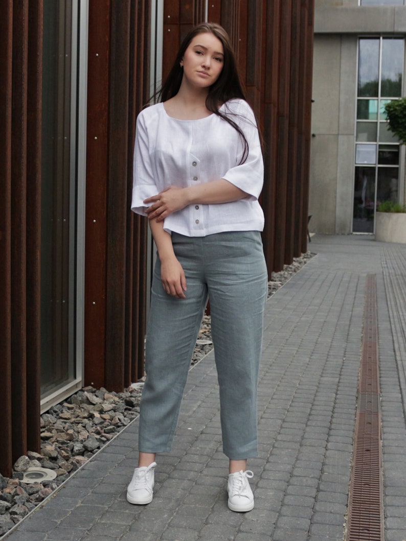 May include: A woman wearing a white linen top with a button closure and light blue linen pants. She is standing in front of a brick wall and is wearing white sneakers.