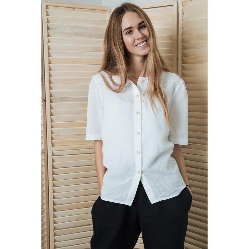 May include: A white linen shirt with a button-up front and short sleeves. The shirt has a subtle dot pattern and is worn with black trousers.