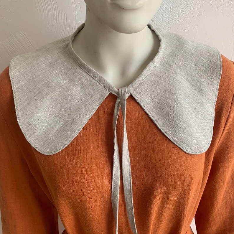 May include: A light gray linen collar with a rounded shape and a tie closure. The collar is worn over a rust-colored linen dress.