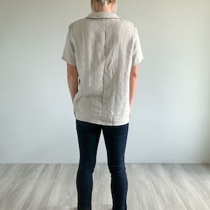 May include: A woman wearing a light beige linen shirt with short sleeves and dark blue jeans. She is standing with her hands in her pockets and wearing white sneakers.