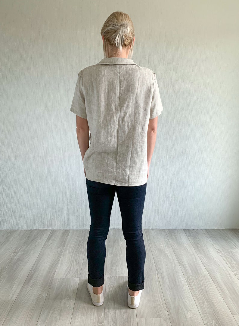May include: A woman wearing a light beige linen shirt with short sleeves and dark blue jeans. She is standing with her hands in her pockets and wearing white sneakers.