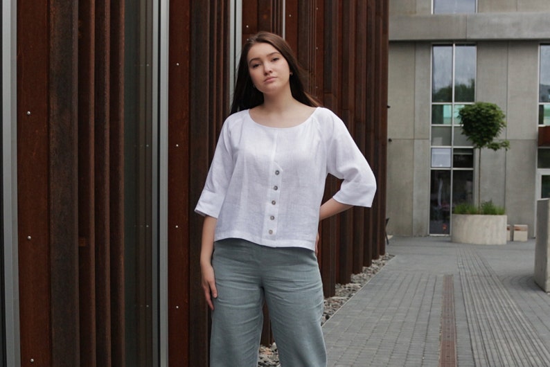 May include: A woman wearing a white linen blouse with a button-up front and a light blue pair of pants.