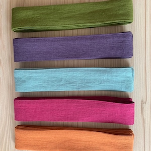 May include: Five fabric strips in different colors: olive green, purple, light blue, burgundy, and orange. The strips are arranged vertically on a light wood surface.