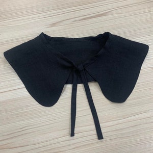 May include: A black fabric collar with a tie at the back. The collar is designed to be worn over a shirt or blouse.