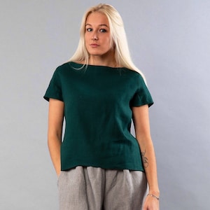 May include: A woman wearing a dark green linen top with short sleeves and a boat neck. She is wearing gray pants.