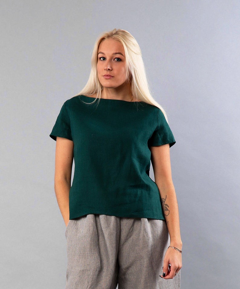 May include: A woman wearing a dark green linen top with short sleeves and a boat neck. She is wearing gray pants.