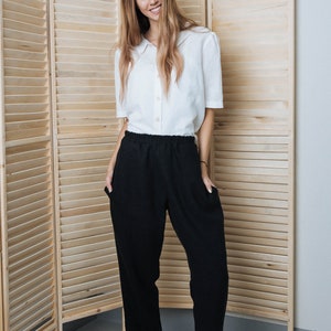 May include: A woman wearing a white collared shirt and black trousers with a relaxed fit. She is standing in front of a wooden room divider and is wearing black and tan shoes.