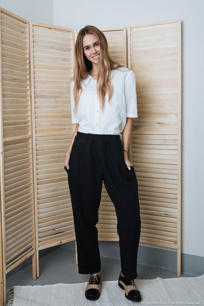 May include: A woman wearing a white collared shirt and black trousers with a relaxed fit. She is standing in front of a wooden room divider and is wearing black and tan shoes.