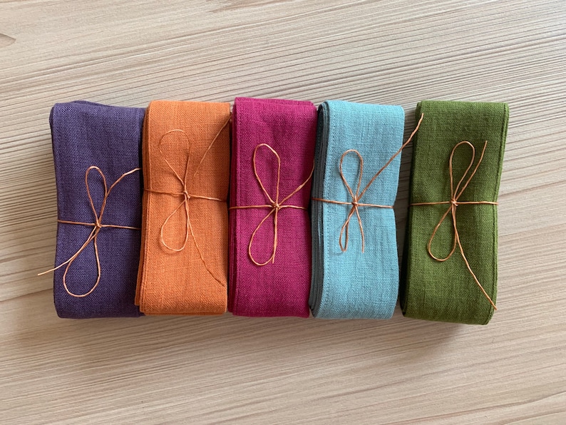 May include: Five rolls of fabric ribbon tied with twine. The ribbons are in shades of purple, orange, pink, blue, and green.