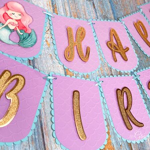 Puede incluir: Bandera morada y azul con letras doradas de purpurina que deletrean "Happy Birthday". La bandera tiene un dise&ntilde;o de sirena con una ilustraci&oacute;n de sirena en la primera bandera.