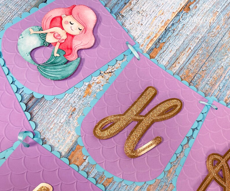 Puede incluir: Bandera morada y azul con una sirena y letras doradas con purpurina que deletrean "Happy". La bandera est&aacute; decorada con un patr&oacute;n de escamas de sirena.