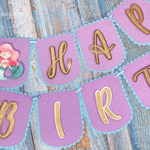 Puede incluir: Bandera morada y azul con letras doradas brillantes que deletrean "Happy Birthday". La bandera presenta una ilustraci&oacute;n de sirena en una de las banderas.