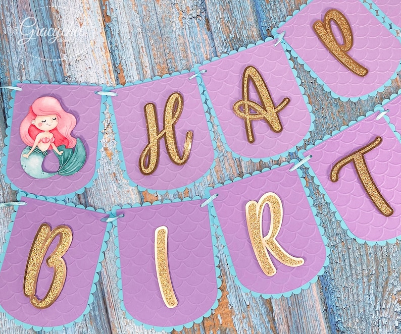 Puede incluir: Bandera morada y azul con letras doradas brillantes que deletrean "Happy Birthday". La bandera presenta una ilustraci&oacute;n de sirena en una de las banderas.
