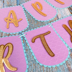 Puede incluir: Una pancarta morada y azul con letras doradas de purpurina que deletrean "Happy" en un tema de sirena.