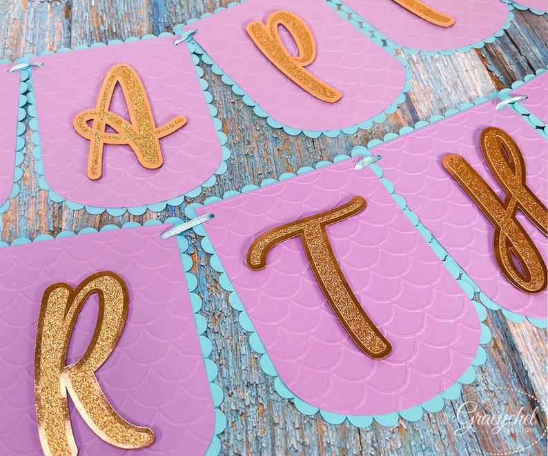 Puede incluir: Una pancarta morada y azul con letras doradas de purpurina que deletrean "Happy" en un tema de sirena.