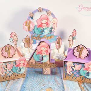 Puede incluir: Un conjunto de decoraciones para fiestas con tem&aacute;tica de sirenas, que incluye un adorno para pastel, cajas de regalo y adornos para cupcakes. Las decoraciones presentan un esquema de colores morado, rosa y dorado con colas de sirena, conchas marinas y el nombre "Valentina".
