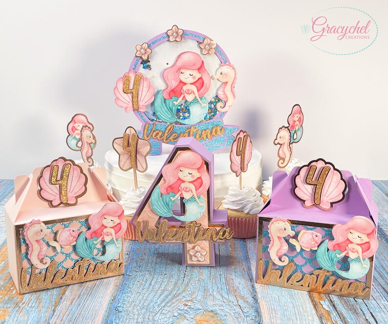 Puede incluir: Un conjunto de decoraciones para fiestas con tem&aacute;tica de sirenas, que incluye un adorno para pastel, cajas de regalo y adornos para cupcakes. Las decoraciones presentan un esquema de colores morado, rosa y dorado con colas de sirena, conchas marinas y el nombre "Valentina".