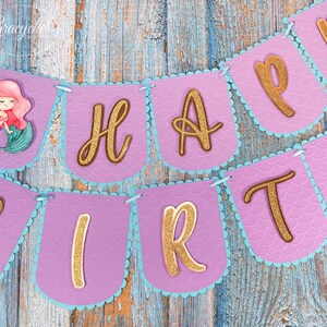 Puede incluir: Bandera morada y azul con letras doradas de purpurina que deletrean "Happy Birthday". Una ilustraci&oacute;n de sirena est&aacute; en el lado izquierdo de la bandera.