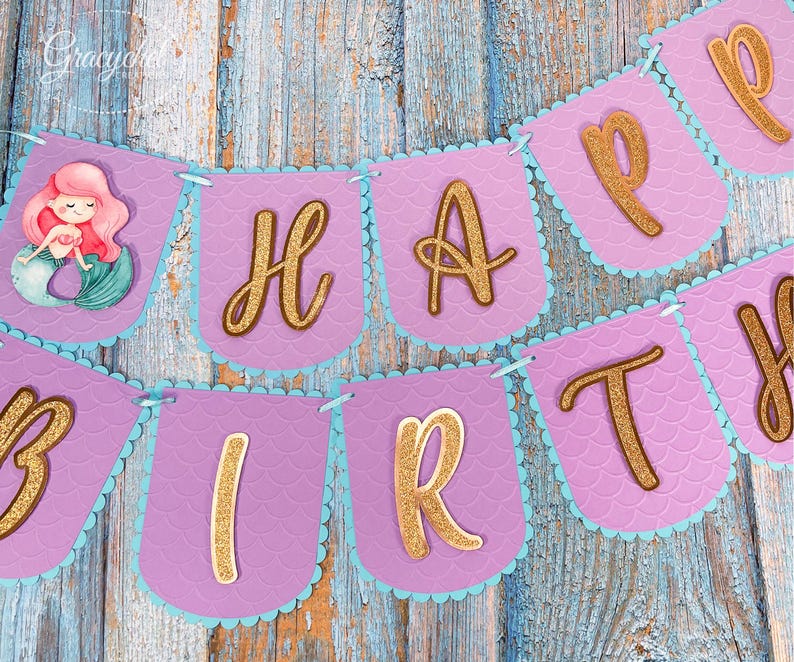 Puede incluir: Bandera morada y azul con letras doradas de purpurina que deletrean "Happy Birthday". Una ilustraci&oacute;n de sirena est&aacute; en el lado izquierdo de la bandera.
