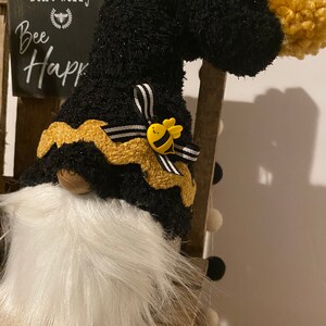 May include: Black and gold gnome with a white beard and a black and white striped bow. The gnome is wearing a yellow bee pin and has a black fuzzy hat with a gold pom-pom. The gnome is sitting on a wooden chair with a sign that says "Don't worry, Bee Happy".