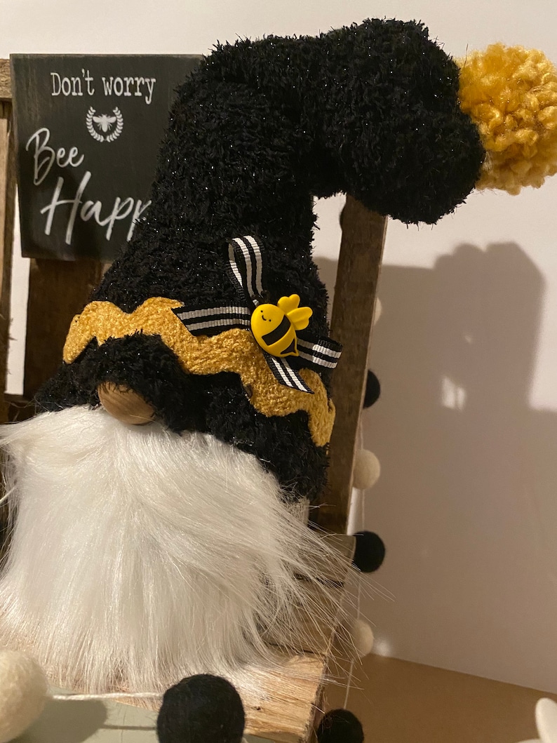 May include: Black and gold gnome with a white beard and a black and white striped bow. The gnome is wearing a yellow bee pin and has a black fuzzy hat with a gold pom-pom. The gnome is sitting on a wooden chair with a sign that says "Don't worry, Bee Happy".