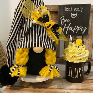 May include: A black and white striped gnome with yellow yarn hair and a bee-themed hat. The gnome is sitting on a wooden base and holding a black mug with the word "SLURP" on it. A sign behind the gnome reads "Don't worry, Bee Happy".