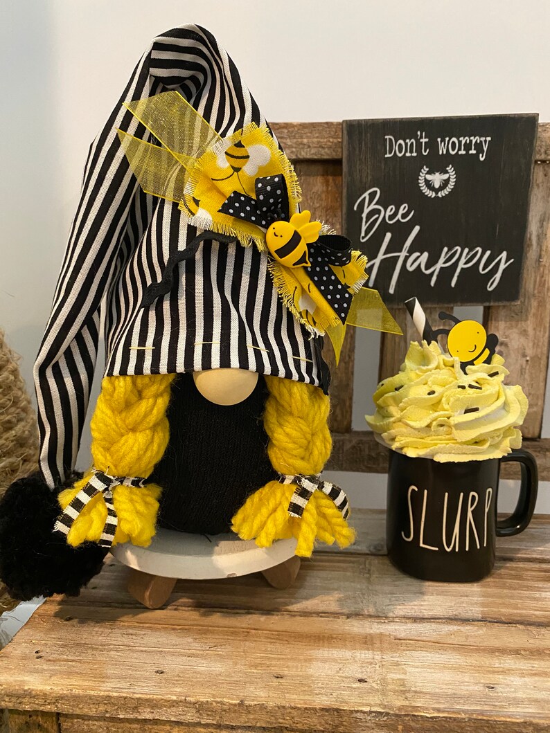 May include: A black and white striped gnome with yellow yarn hair and a bee-themed hat. The gnome is sitting on a wooden base and holding a black mug with the word "SLURP" on it. A sign behind the gnome reads "Don't worry, Bee Happy".