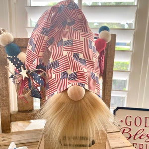 May include: Patriotic gnome with a red, white, and blue American flag patterned hat. The gnome has a blonde beard and is sitting on a wooden box.