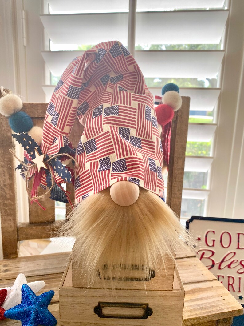 May include: Patriotic gnome with a red, white, and blue American flag patterned hat. The gnome has a blonde beard and is sitting on a wooden box.
