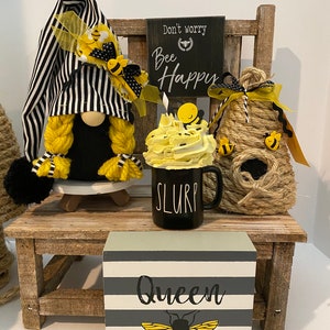May include: A black and white striped gnome with yellow yarn hair and a yellow bee hat sits on a wooden chair with a black sign that says "Don't worry, Bee Happy". A yellow and white bee-themed mug with the word "SLURP" on it sits in front of the gnome. A small wooden block with gray and white stripes and a bee graphic says "Queen".