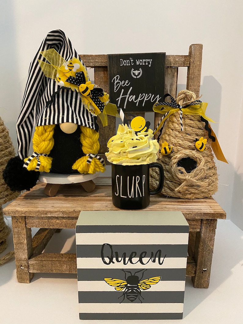 May include: A black and white striped gnome with yellow yarn hair and a yellow bee hat sits on a wooden chair with a black sign that says "Don't worry, Bee Happy". A yellow and white bee-themed mug with the word "SLURP" on it sits in front of the gnome. A small wooden block with gray and white stripes and a bee graphic says "Queen".