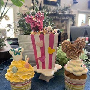 May include: A decorative display featuring three cupcakes and a popcorn container. The cupcakes have colourful frosting and Mickey Mouse-themed decorations. The popcorn container is pink and white with Mickey Mouse silhouettes.
