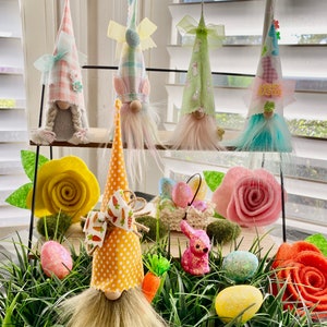 May include: Four Easter gnomes with different colored hats and beards, sitting on a shelf with a pink and yellow felt flower, a basket of Easter eggs, a pink bunny, and a carrot. The gnomes are wearing hats with different patterns, including plaid, polka dots, and floral prints. The gnomes are surrounded by green grass and Easter eggs.