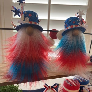 May include: Two gnomes with red, white, and blue fur, wearing red, white, and blue hats with stars and glitter. The gnomes are decorated for the Fourth of July.